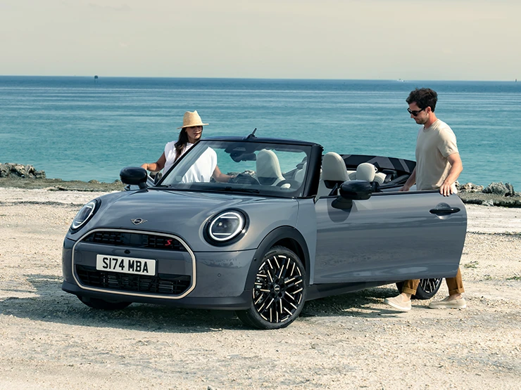 Prikaz lijevog prednjeg kraja MINI Cooper Cabrio modela u Copper Grey boji na plaži s dvije osobe pored njega.