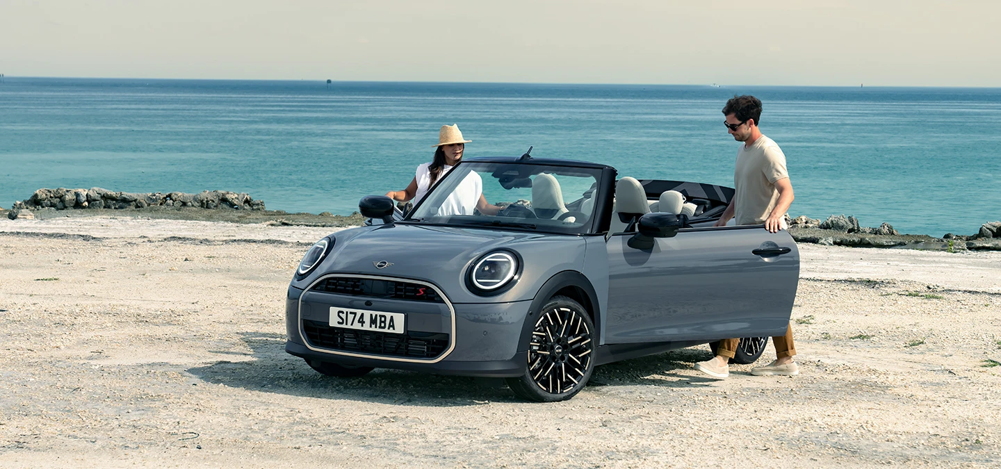 Prikaz lijevog prednjeg kraja MINI Cooper Cabrio modela u Copper Grey boji na plaži s dvije osobe pored njega.