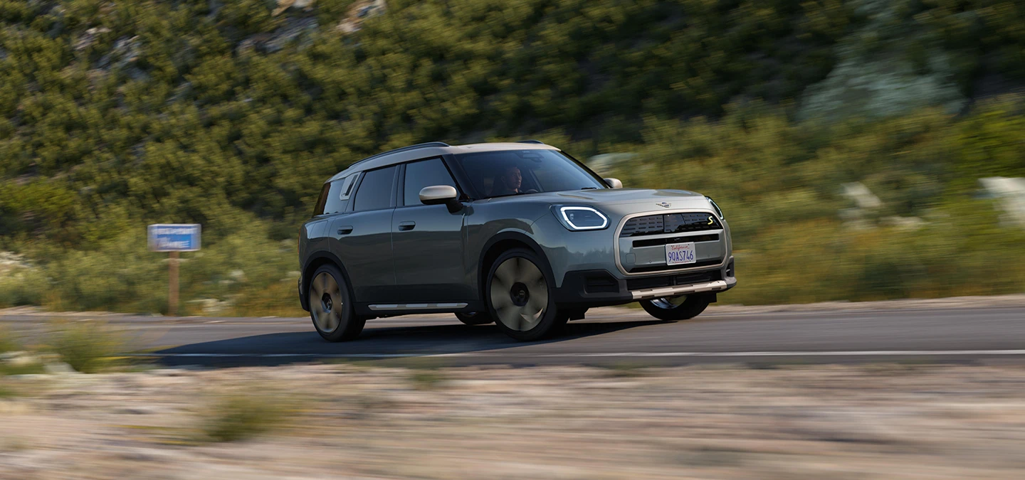 Bočni prikaz stražnjeg kraja potpuno električnog MINI Countryman modela u Smokey Green boji i krova u Vibrant Silver Roof boji u offroad vožnji.