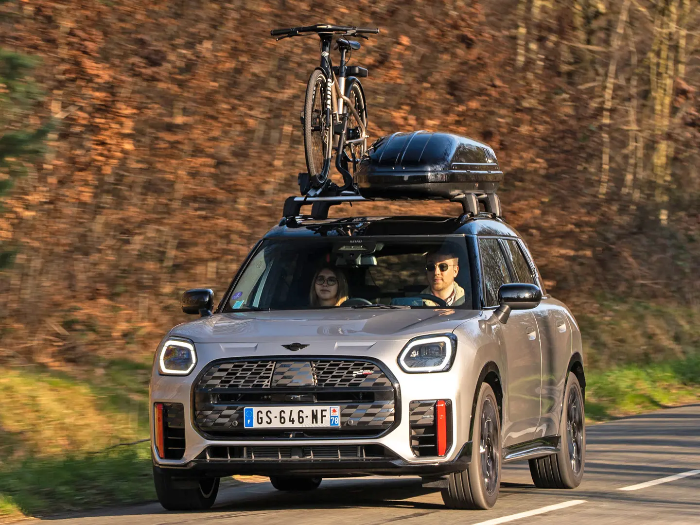 MINI Countryman Cooper u Silver boji na planinskoj cesti s MINI krovnim nosačem, MINI krovnom kutijom, MINI krovnim nosačem bicikla s jednim biciklom plus MINI John Cooper Works krilo - prikaz prednjeg kraja. 