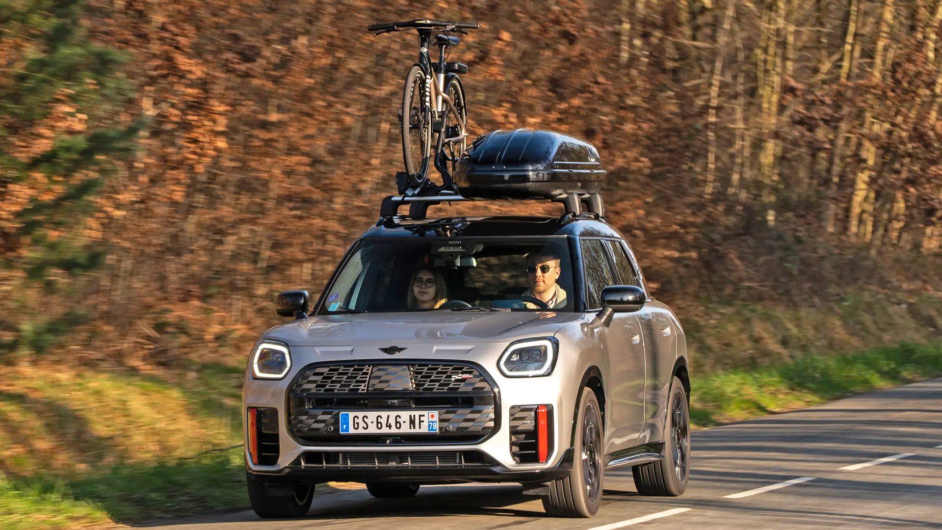 MINI Countryman Cooper u Silver boji na planinskoj cesti s MINI krovnim nosačem, MINI krovnom kutijom, MINI krovnim nosačem bicikla s jednim biciklom plus MINI John Cooper Works krilo - prikaz prednjeg kraja. 