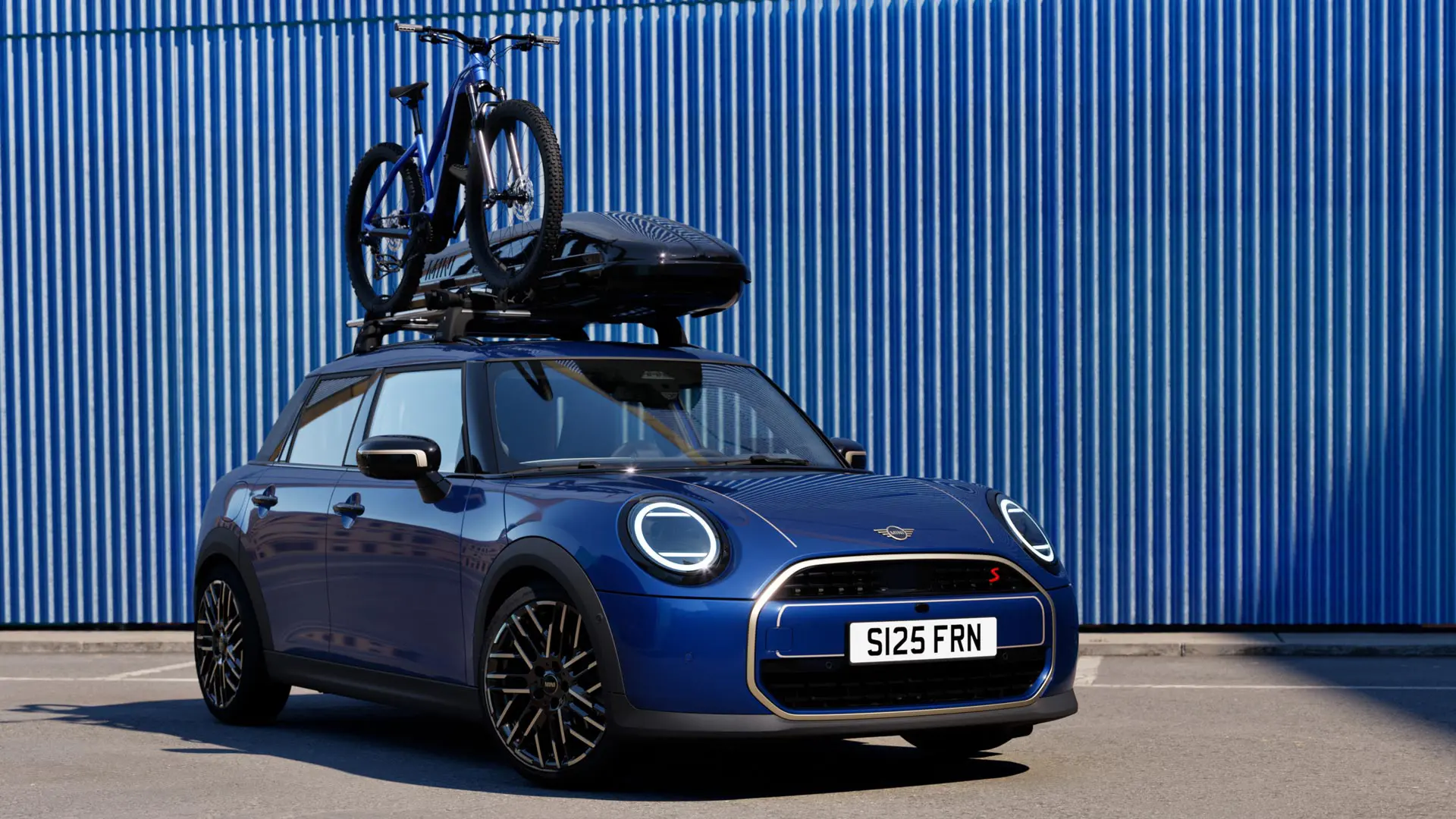Prikaz prednjeg kraja na MINI Aceman u Indigo Sunset Blue boji s MINI krovnim nosačem za bicikle, s brdskim biciklom i MINI Roof Box 320.
