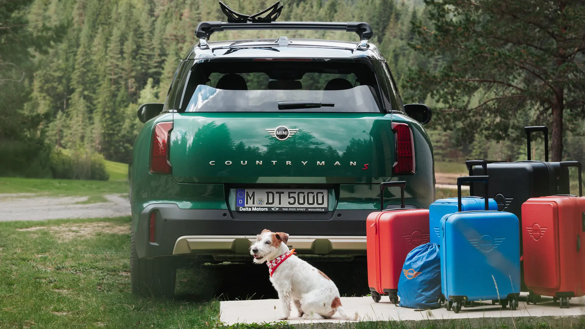 MINI Countryman u zelenoj boji s raznim predmetima prtljage dostupne u MINI Lifestyle trgovini, stoji iza vozila zajedno s Jack Russel psom.