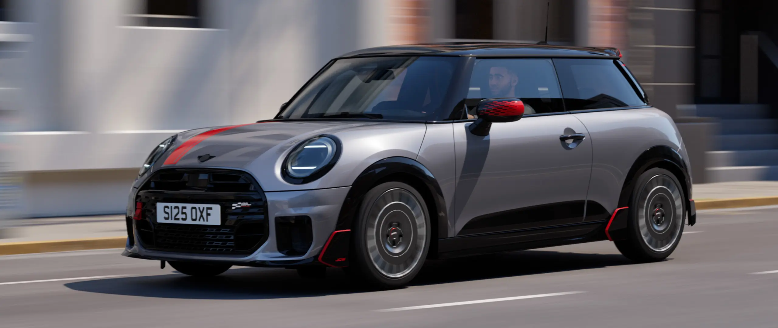 MINI Cooper 3-vrata u Melting Silver boji s John Cooper Works kućištima retrovizora i naljepnicom poklopca motora u Chili Red boji, krovni spojler i krilo u Black boji, vožnja gradskom ulicom - pogled sprijeda.